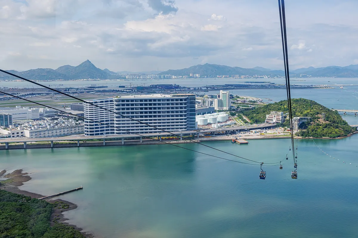 Looking down from a cabin over the green water of Tung Chung Bay to a white building, a wooded headland and the airport, with cabins on the cable below