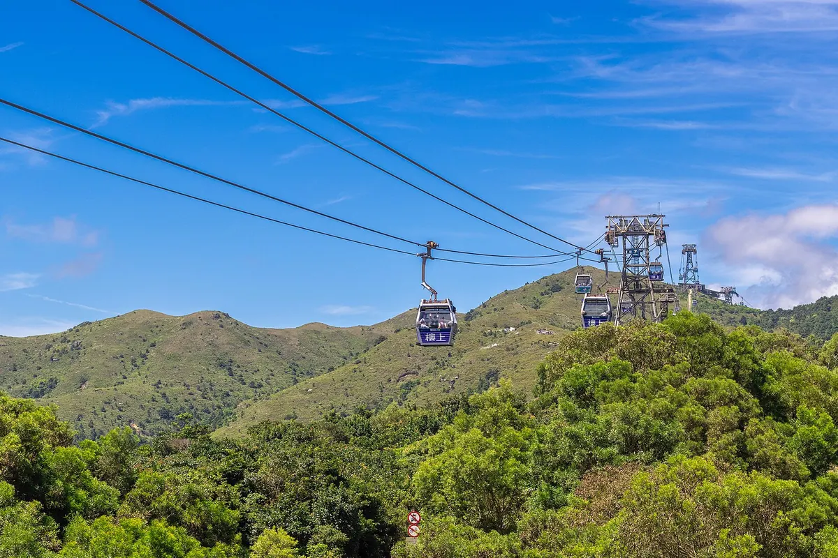 A blue cabin hangs from the cable above green hills, with a steel tower and more cabins climbing away under a bright blue sky